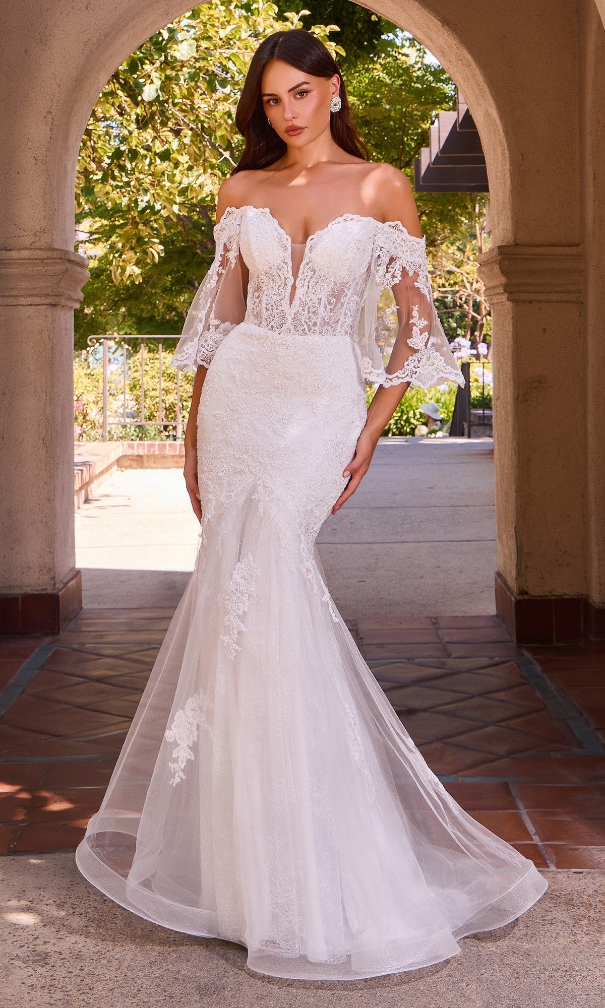 A woman stands outdoors under an archway, wearing the Ladivine CD659W long wedding dress in off white—a fitted off-the-shoulder gown with sheer sleeves and a graceful mermaid skirt.